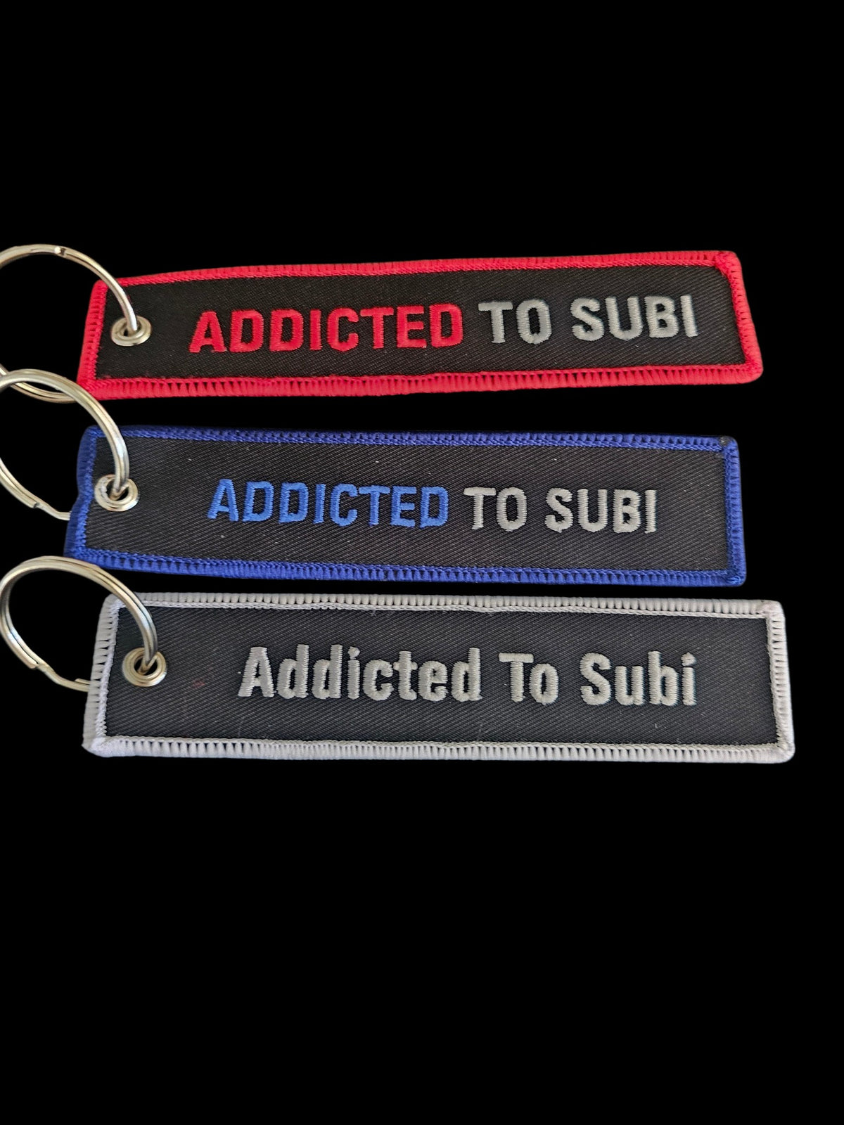 Three Addicted To Subi embroidered fabric key tags with metal rings in red, blue, and white stitching on black.